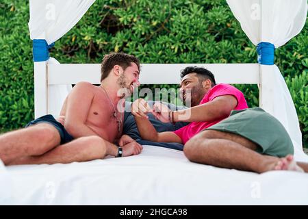 Portrait eines homosexuellen Ehepaares von zwei Männern, das auf einem Strandbett in einem Strandclub in cancun liegt, während er spielt und lacht, lgbti-Konzept Stockfoto