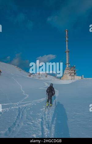 Person Skitouren bergauf mit großen Beton-Fernseher und Radio-Antenne auf der Skipiste Krvavec in der Abendsonne. Stockfoto