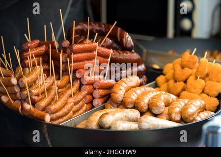 Köstliche gegrillte und gebratene Würstchen und andere Speisen mit Stöcken. Street Food auf dem lokalen Markt des traditionellen Festivals. Lebensmittelhändler auf der Straße Stockfoto