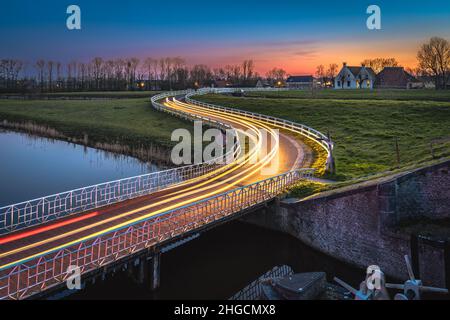 Geschwungene asphalanische Landstraße in holländischer Landschaft mit weißem Zaun, der zu einem alten Bauernhaus in Holland führt. Stockfoto