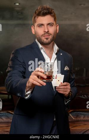 Selbstbewusster junger Pokerspieler mit zwei Assen und einem Glas Whiskey im Casino Stockfoto