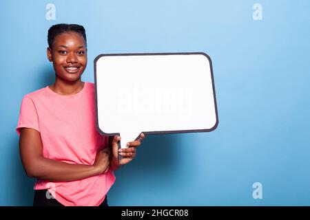 Porträt einer afroamerikanischen jungen Frau, die lächelt, während sie im Studio mit blauem Hintergrund ein weißes Quizbanner hält. Werbekonzept. Whiteboard-Konzept Stockfoto