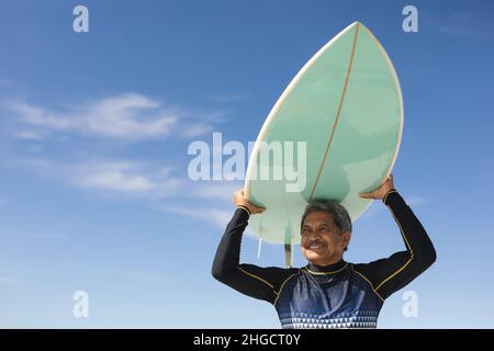 Blick aus der unteren Perspektive auf den Senioren, der am sonnigen Strand gegen den blauen Himmel Surfbrett über den Kopf trägt Stockfoto