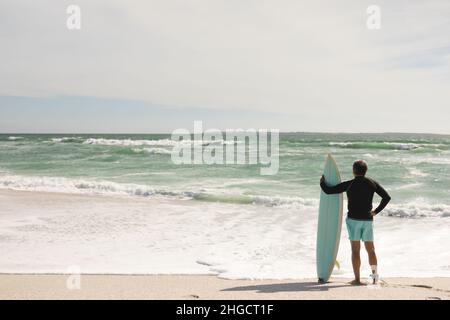 Der erfahrene Biracial-Mann, der an sonnigen Tagen am Strand an der Küste am Surfbrett steht, hält die ganze Länge Stockfoto