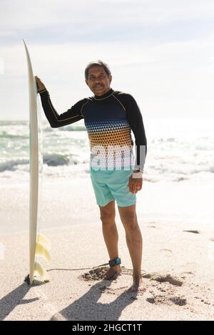 In voller Länge Porträt eines älteren Biracial-Mannes, der am Surfbrett am sonnigen Strand gegen den Himmel steht Stockfoto