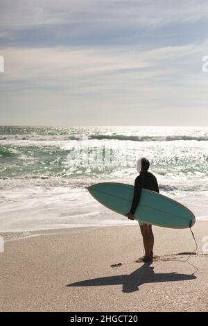 Biracial Senior Mann trägt Surfbrett Blick auf die plätschernden Wellen im Meer gegen den Himmel an sonnigen Tag Stockfoto