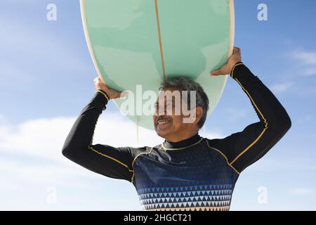 Blick aus der unteren Perspektive auf einen lächelnden Senioren, der am sonnigen Strand Surfbrett mit dem Kopf gegen den Himmel trägt Stockfoto