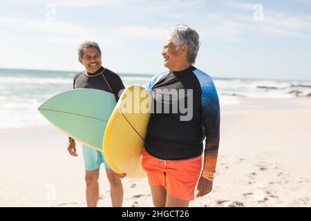 Lächelndes, multirassisches Seniorenpaar, das sich an sonnigen Tagen mit Surfbrettern an Land ansieht Stockfoto