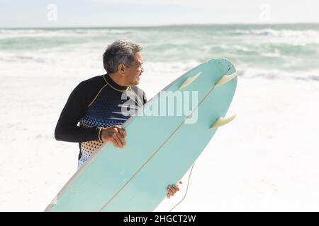 Biracial Senior Mann trägt blaues Surfbrett an der Küste, während Blick auf das Meer an sonnigen Tagen Stockfoto