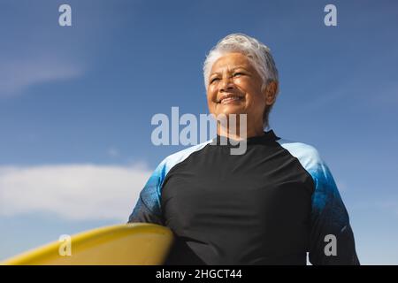 Aus der Perspektive der lächelnden älteren Birazialfrau, die an sonnigen Tagen Surfbrett gegen den blauen Himmel trägt Stockfoto