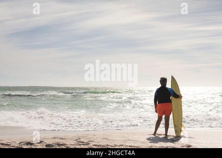 Lange Rückansicht einer älteren Frau mit dem Surfbrett, die vom sonnigen Strand aus den Horizont blickt Stockfoto