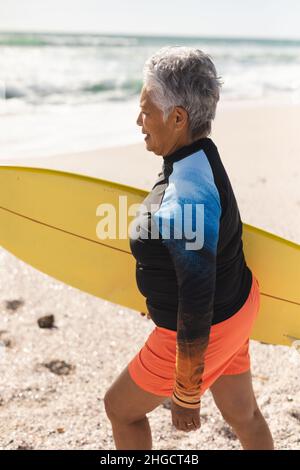 Seitenansicht einer älteren Frau im Neoprenanzug, die mit gelbem Surfbrett am sonnigen Strand spazierengeht Stockfoto