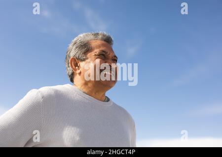 Blick aus der Ferne auf einen lächelnden Senioren, der an sonnigen Tagen vor dem blauen Himmel auf den Strand blickt Stockfoto