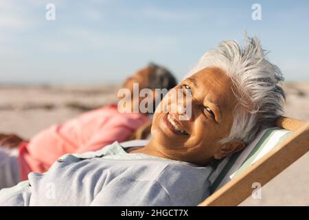Porträt einer lächelnden älteren Birazialfrau, die sich an sonnigen Tagen mit einem Mann am Strand auf einem Stuhl entspannt Stockfoto