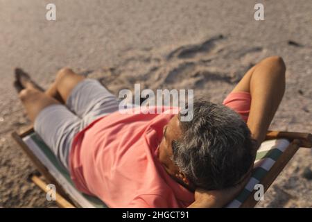 Blick aus der VogelPerspektive auf einen pensionierten älteren Birazialmann, der sich mit den Händen hinter dem Kopf auf dem Stuhl am Strand entspannt Stockfoto