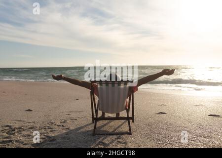 Pensionierter älterer Birazialmann mit ausgestreckten Armen, der während des Sonnenuntergangs auf einem Klappstuhl am Strand sitzt Stockfoto