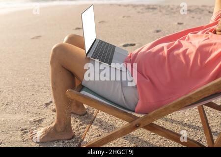 Niedriger Teil eines entspannten älteren Birazialmannes, der mit einem Laptop auf einem Klappstuhl am Strand sitzt Stockfoto