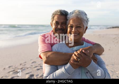 Porträt eines liebevollen älteren Birazialmannes, der die Frau während des Sonnenuntergangs am Strand von hinten umarmt Stockfoto