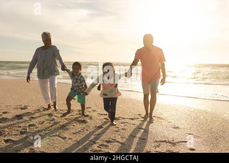 Multirassische Enkelkinder halten sich die Hände, während die Großeltern während des Sonnenuntergangs am Strand spazieren Stockfoto