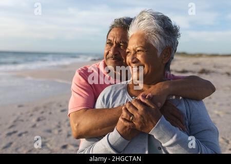 Nachdenkliches, mehrrassiges Paar, das sich umarmte, während es bei Sonnenuntergang auf den Strand schaute Stockfoto