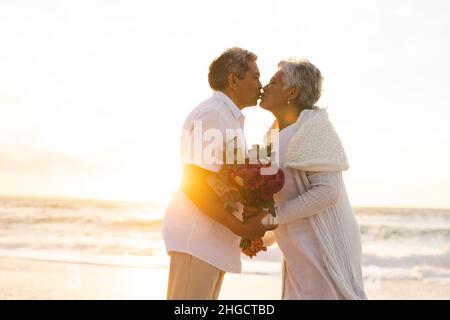 Seitenansicht eines mehrrassigen älteren Ehepaares, das sich während der Hochzeitszeremonie am Strand küsst Stockfoto