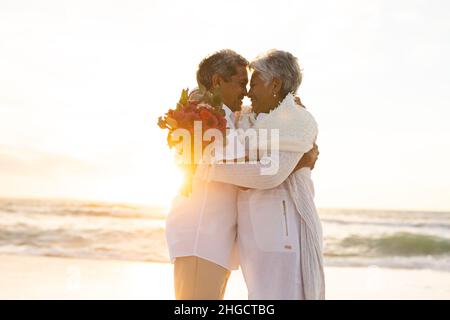 Ein glückliches, frisch verheiratetes, mehrrassiges Ehepaar umarmt sich, während es während des Sonnenuntergangs am Strand einen Blumenstrauß hält Stockfoto