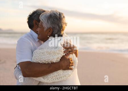 Glückliche, frisch vermählte, multirassische ältere Braut und Bräutigam umarmen sich am Strand während des Sonnenuntergangs Stockfoto