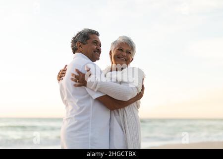 Lächelndes, frisch verheiratetes, multirassisches Paar, das sich umarmte, während es während des Sonnenuntergangs auf den Strand schaute Stockfoto