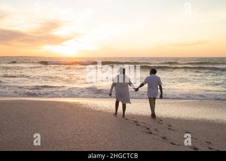 Rückansicht eines mehrrassischen Ehepaares, das während des Sonnenuntergangs am Strand herumläuft und die Hände hält Stockfoto