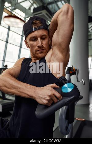 Ein junger Mann benutzt und genießt die Schlagmassagepistole, um sich zu entspannen und die Schmerzen im Körper und in den Muskeln im Fitnessstudio zu lindern. Stockfoto