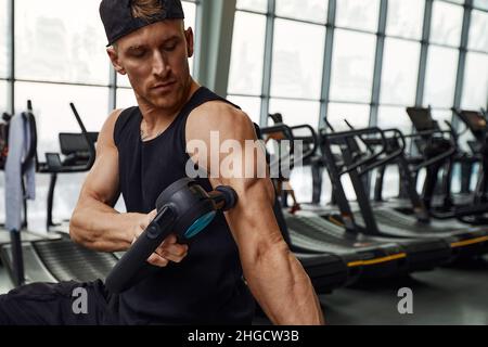 Ein junger Mann benutzt und genießt die Schlagmassagepistole, um sich zu entspannen und die Schmerzen im Körper und in den Muskeln im Fitnessstudio zu lindern. Stockfoto
