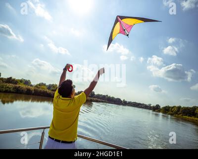 Junger Mann steht auf der Terrasse des schwimmenden Hauses und beginnt, hellen Drachen in den Himmel zu fliegen, Foto von hinten Stockfoto