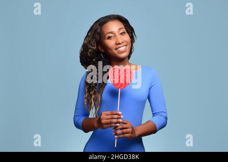 Schönes schwarzes Mädchen in einem blauen Kleid lächelt und hält einen großen herzförmigen Lollipop auf einem Stock. Stockfoto