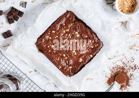 Schokoladen-Fudge-Brownies mit Haselnussbröseln oben, auf einem Tablett mit Kakaopulver, Schokoladenbröseln auf weißem, rustikalem Hintergrund. Stockfoto
