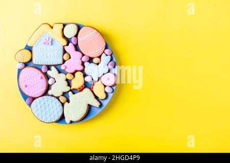 Frohe Ostern. Mehrfarbiger Pastell-osterkekse-Teller auf gelbem Hintergrund, verschiedene Lebkuchenglasierte Kekse, flaches Lay, Blick von oben, Freiraum für Grußtext, Banner, Flyer, Gutschein. Stockfoto