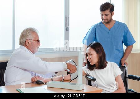 kaukasischer leitender Arzt mit Sphygmomanometer Überprüfung des Blutdrucks und berät junge asiatische Patientin Frau im Krankenhaus. Gesundheitsversorgung und Sanitäter Stockfoto