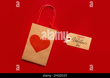 Draufsicht auf Einkaufstasche mit rotem Herz und braunem Sale-Tag mit Text Happy Valentine's day auf rotem Hintergrund. Konzept für die Valentinstag-Feier Stockfoto