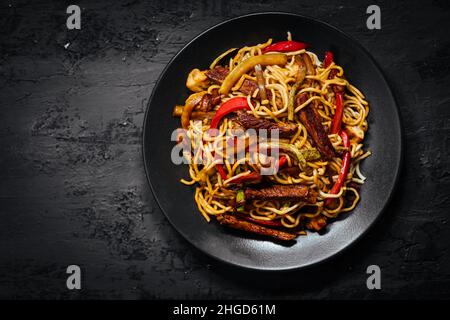 Wok mit Nudeln, Paprika, Pilzen, putenfleisch und verschiedenen Gemüsesorten und Sesamsamen in einer blauen Schüssel auf dunklem Hintergrund Stockfoto