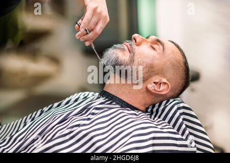 Barbier, der in einem professionellen Friseursalon oder Salon einen männlichen Kundenbart mittleren Alters mit einer Schere in einer Nahaufnahme auf der Hand und dem Pusto trimmen konnte Stockfoto