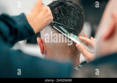 Profi-Friseur mit Schere und Kamm, um ein mans Haar in eine moderne trendige V-förmige Frisur in einer Nahaufnahme Rückansicht auf seine Hände und die zu schneiden Stockfoto