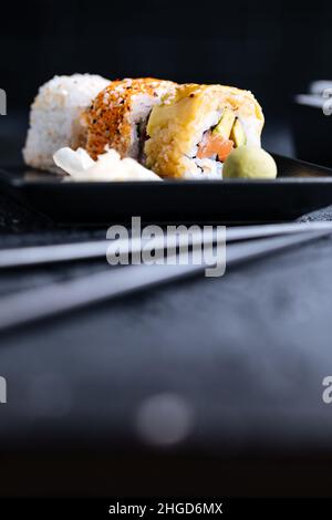 Verschiedene Sushi-Rollen auf dunklem Hintergrund mit Essstäbchen, Wasabi und Sojasauce. Traditionelles japanisches Street Food Stockfoto