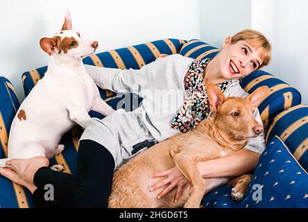 Fröhliche Besitzerin, die auf dem blauen Sofa liegt und zwei niedliche Pharaoh Hound Hunde im hellen Wohnzimmer zu Hause umarmt Stockfoto