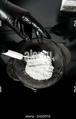 Weibliche Hände in medizinischen Handschuhen halten eine Schüssel mit weißem Pulver. Kaolin Tonerde weißes Pulver kosmetische Qualität für Gesichtsmaske und Spa-Behandlungen. Vertikales Foto Stockfoto