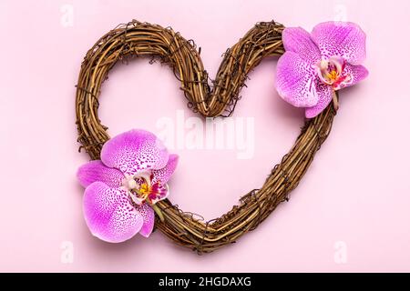 Herzförmiger Rattan-Kranz mit tropischen Orchideenblüten auf rosa Hintergrund Draufsicht Flat Lay Holiday Card Happy Valentine's day, love con Stockfoto
