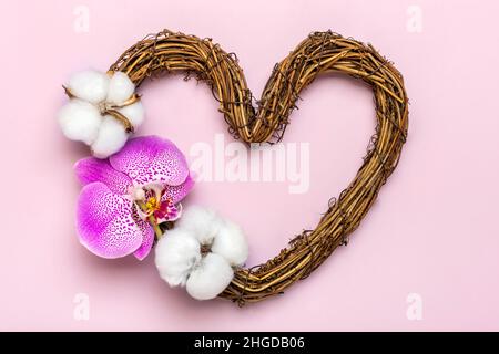 Herzförmiger Rattan-Kranz mit tropischen Orchideenblüten, Baumwolle auf rosa Hintergrund Draufsicht Flat Lay Holiday Card Happy Valentine's day, Stockfoto