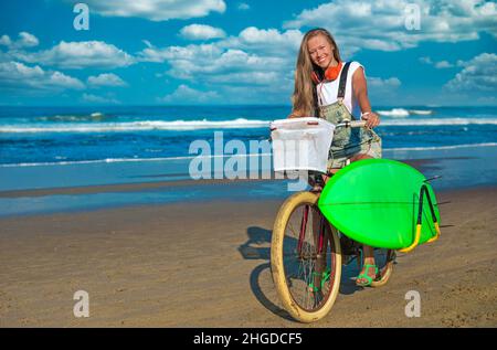 Junge Frau mit Surfbrett und Fahrrad am Strand. Stockfoto