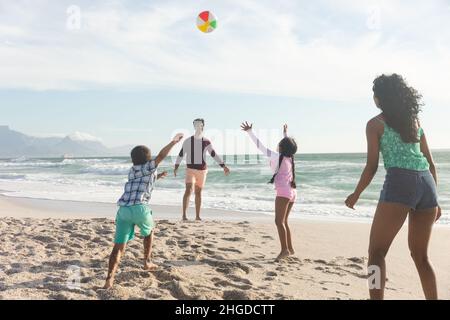 Glückliche multirassische Familie, die an sonnigen Tagen am Strand gegen den Himmel mit Ball auf Sand spielt Stockfoto