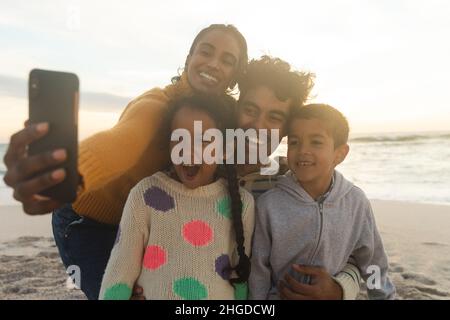 Glückliche Birazialfrau, die während des Sonnenuntergangs ein Selfie der Familie über das Smartphone am Strand macht Stockfoto