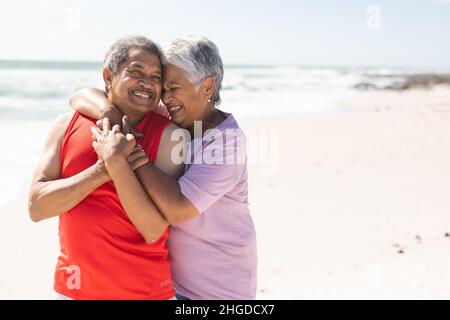 Fröhliche ältere Frau aus der Birazialzeit umarmt den Mann von hinten am Strand gegen das Meer an einem sonnigen Tag Stockfoto