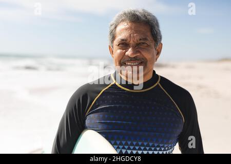 Porträt eines lächelnden Senioren aus dem Ruhestand, der an einem sonnigen Tag mit Surfbrett am Strand steht Stockfoto